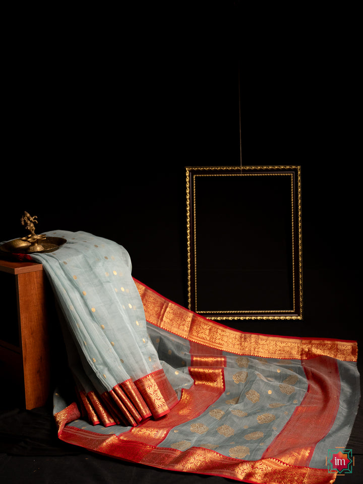 Elegant grey silk saree is pleated and displayed upon a bench with a ganapati statue upon it and with a black background.