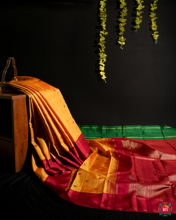 Yellow Red Ganga Jamuna Kanjivaram silk saree is displayed on the floor with black background and yellow flowers hanging above.